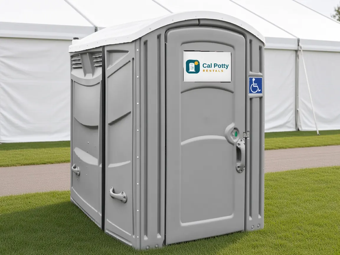 A grey ADA-accessible portable restroom with a wheelchair sign on a lawn in front of large white tents
