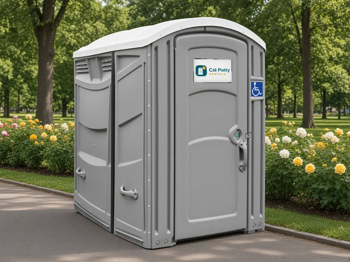 A grey ADA-accessible portable restroom with a wheelchair sign on a park path beside rose bushes and trees